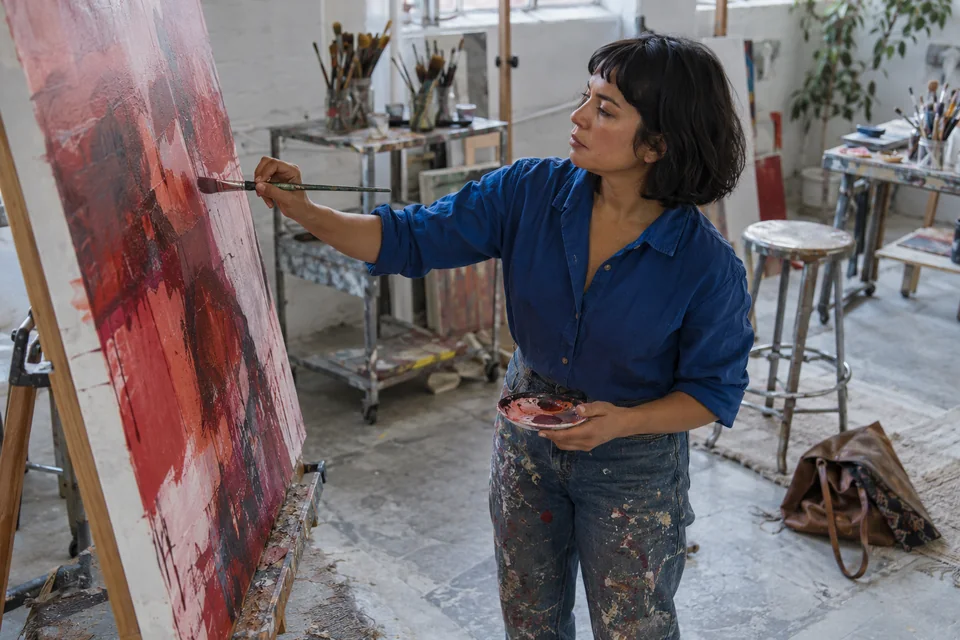 Latina adulta trabaja en un atelier creativo con superficies azules y detalles rojos, vista desde arriba. Imagen editorial generada con IA; personas ficticias.