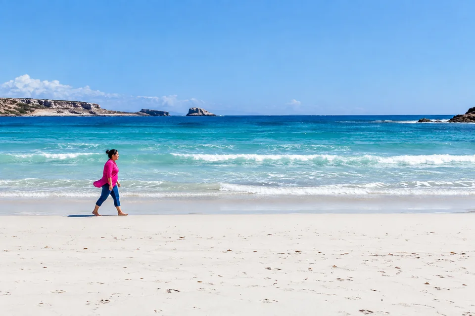 Mujer latina con camisa fucsia camina en una costa amplia; el mar y el cielo dominan la composición. Imagen editorial generada con IA; personas ficticias.