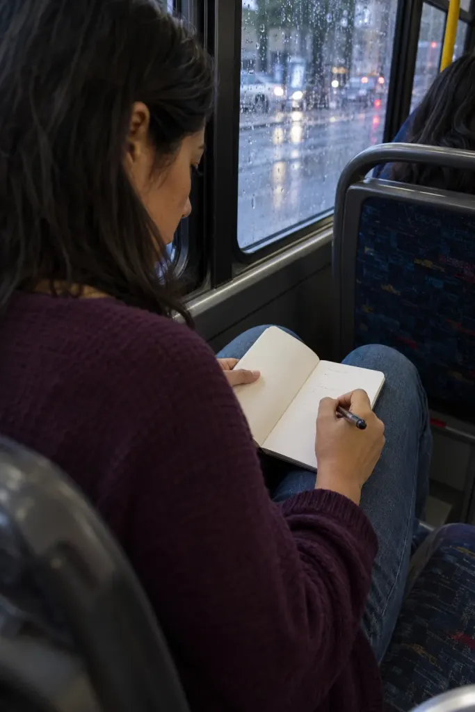 Una mujer escribe en una libreta sobre sus piernas mientras viaja junto a una ventana de autobús.
