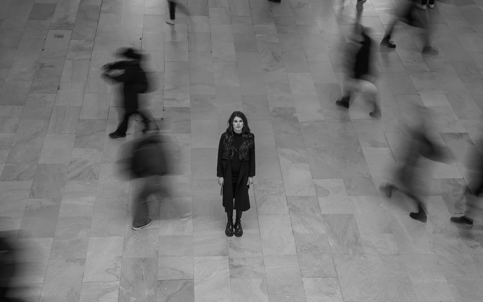 Fotografía en blanco y negro de una mujer de pie, inmóvil, mientras la gente pasa a su alrededor
