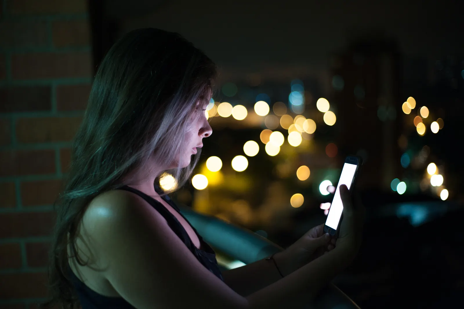 Mujer joven mira la pantalla de su celular de noche en un balcón con luces de la ciudad
