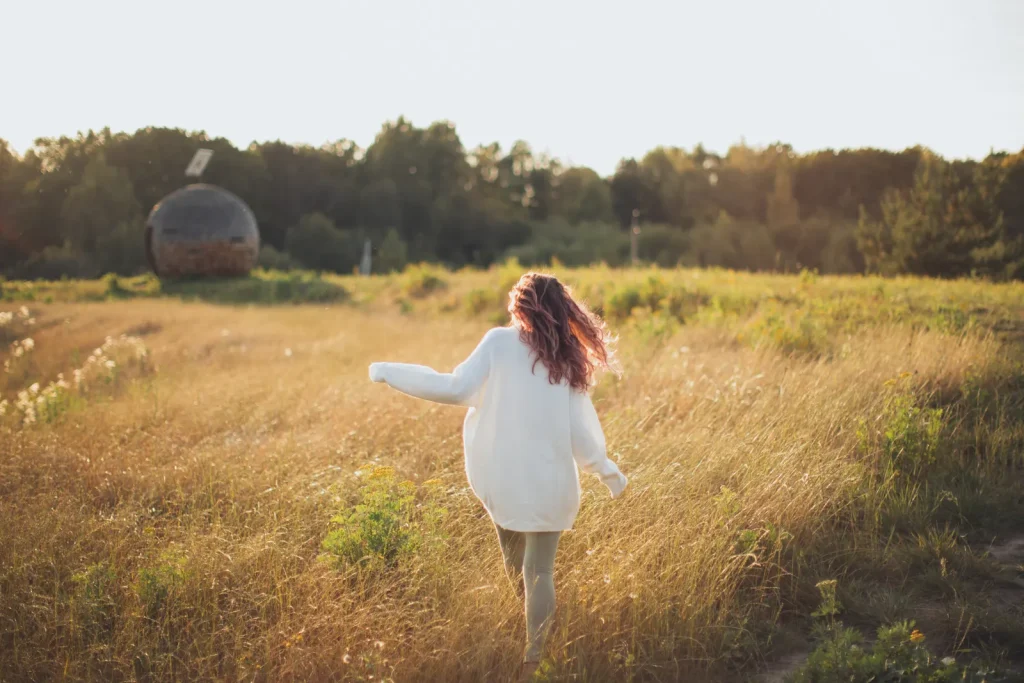 Mujer de espaldas con suéter blanco camina por un campo de hierba dorada al atardecer