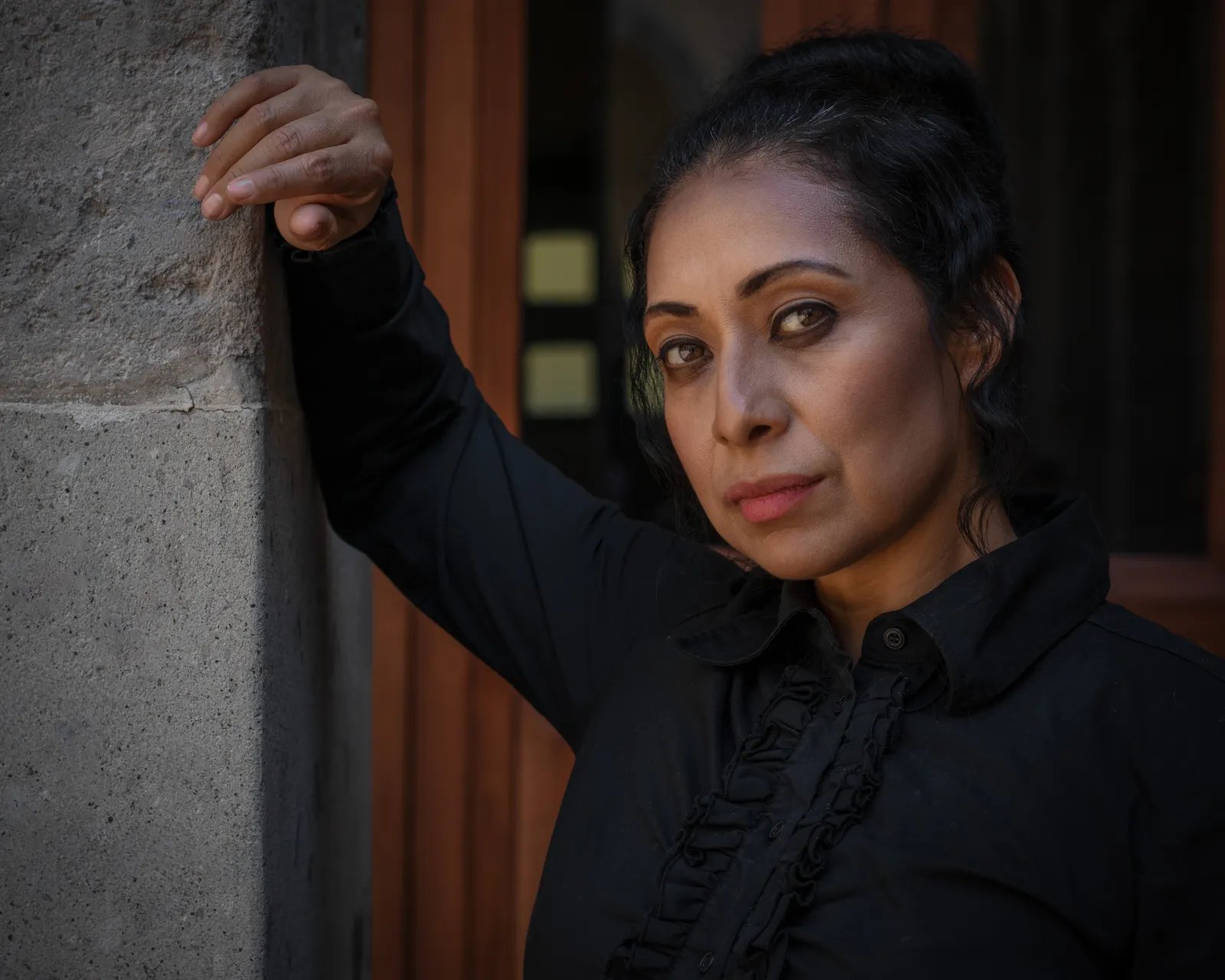 Mujer de cabello recogido y blusa negra mira a la cámara apoyada en un muro de piedra