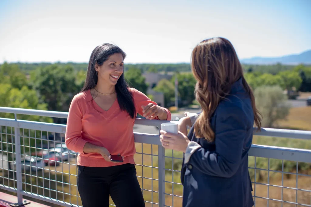 Dos mujeres conversan de pie en una terraza soleada con árboles al fondo