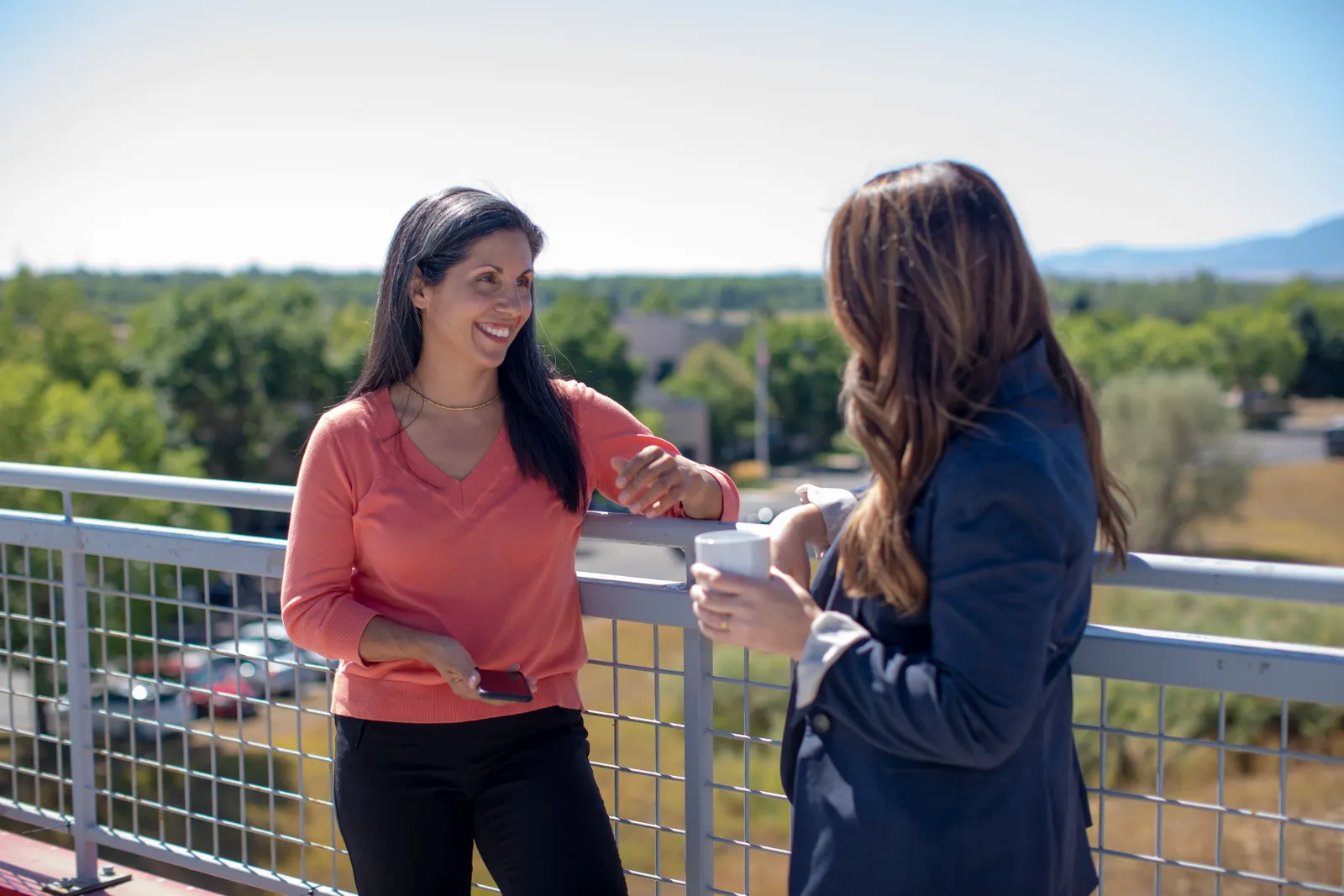 Dos mujeres conversan de pie en una terraza soleada con árboles al fondo