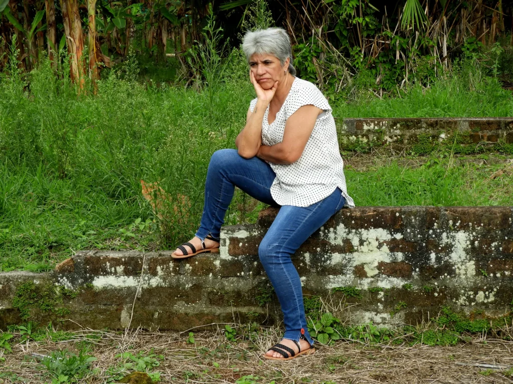 Mujer de cabello canoso apoya la barbilla en la mano, pensativa, sentada sobre un muro en un jardín
