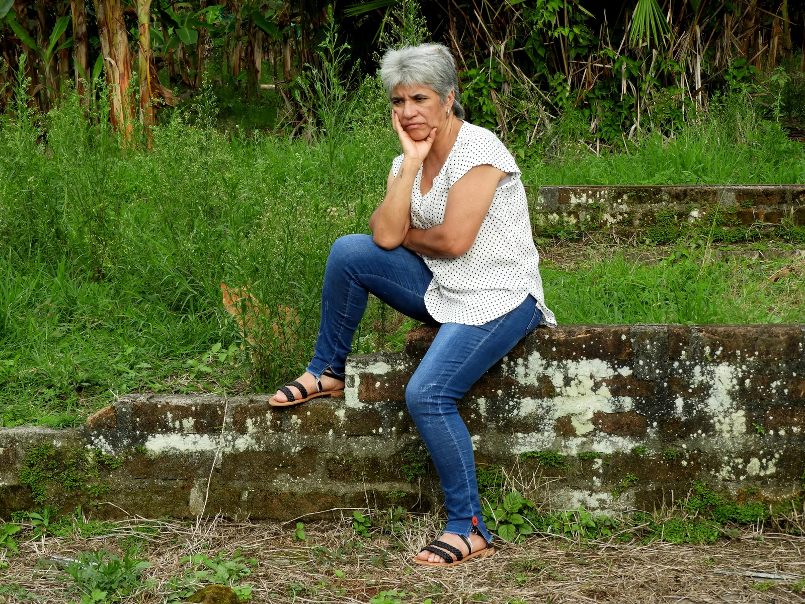 Mujer de cabello canoso apoya la barbilla en la mano, pensativa, sentada sobre un muro en un jardín