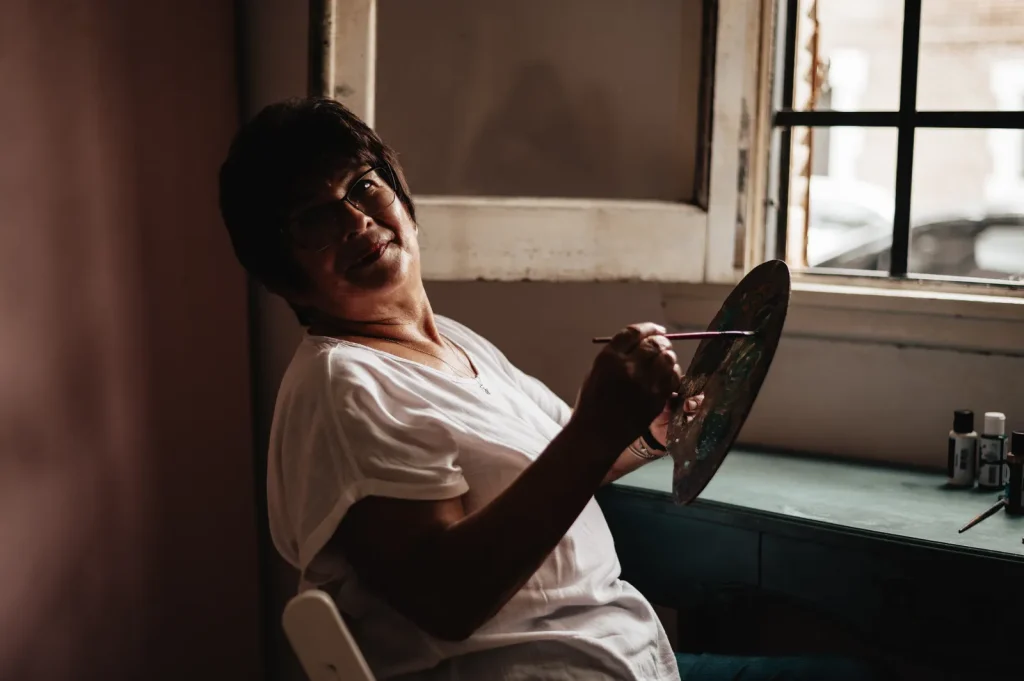Mujer ríe mientras pinta con paleta y pincel junto a una ventana con luz natural