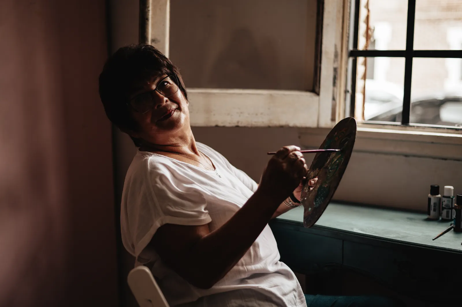 Mujer ríe mientras pinta con paleta y pincel junto a una ventana con luz natural