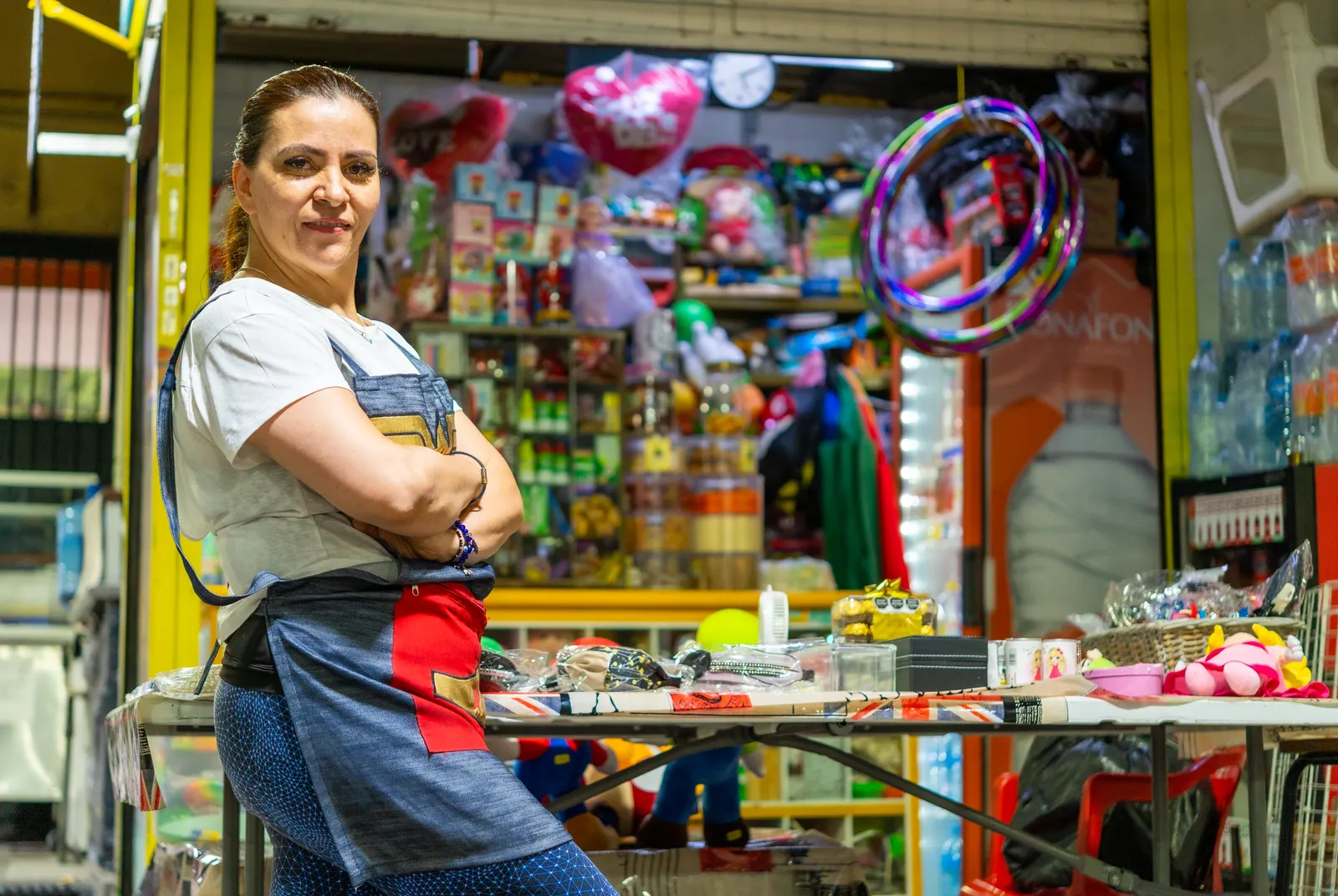 Mujer con delantal y brazos cruzados posa con orgullo frente a su puesto de juguetes en un mercado