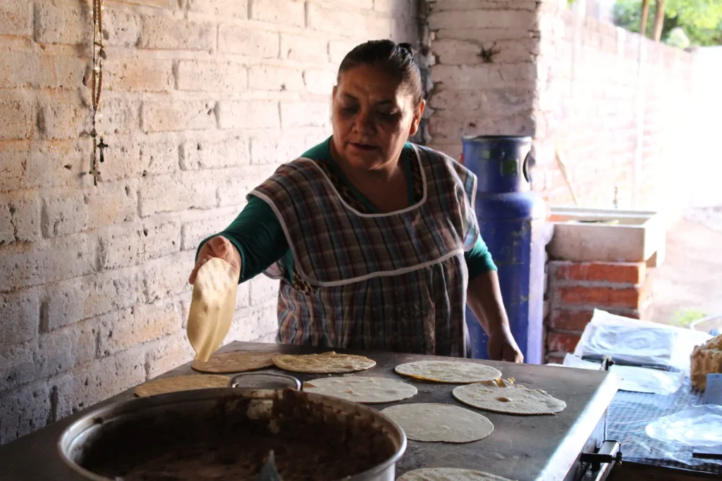 Mujer con mandil prepara tortillas a mano sobre un comal en una cocina con muros de ladrillo