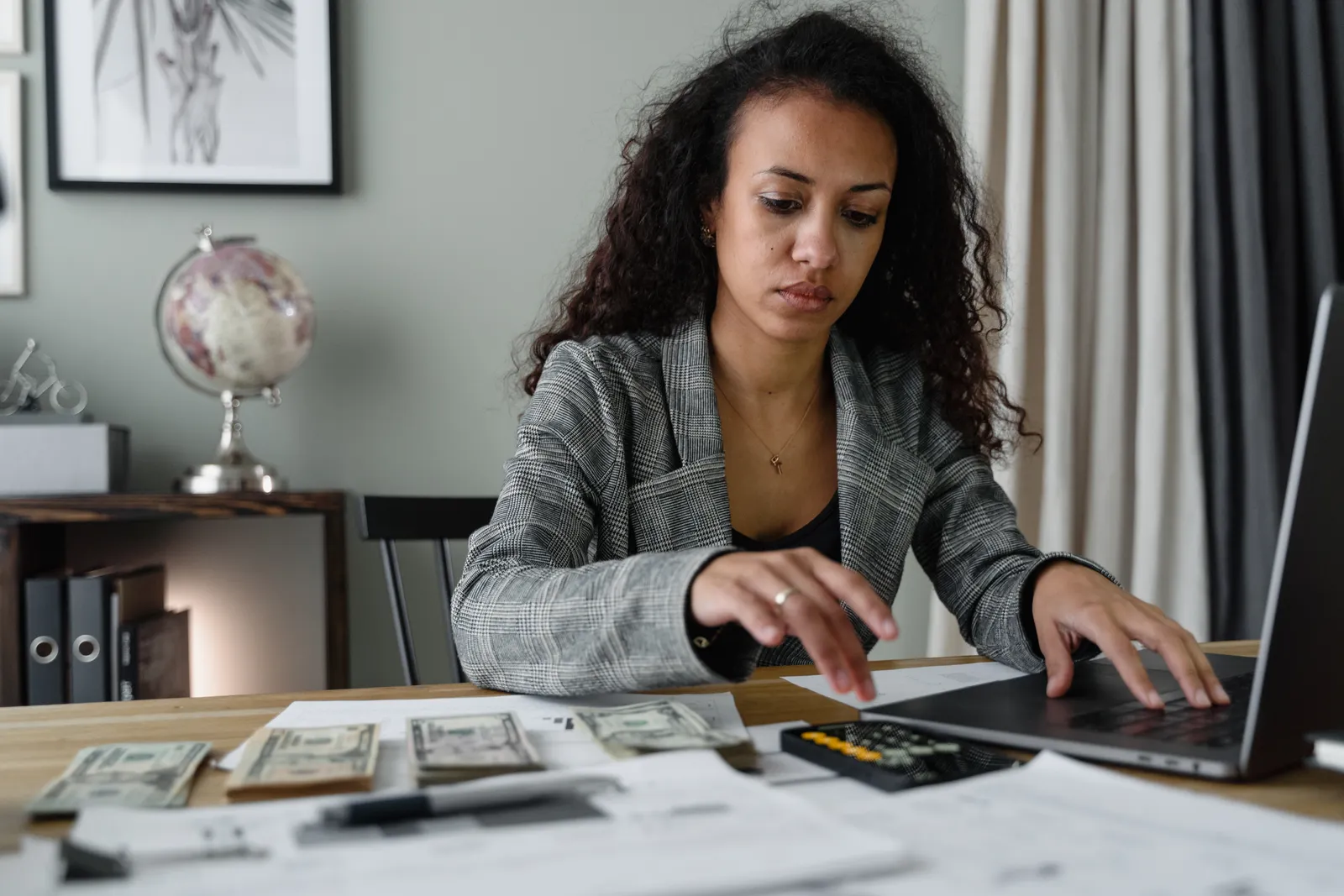 Mujer de cabello rizado revisa sus finanzas en una computadora portátil con documentos y billetes sobre el escritorio