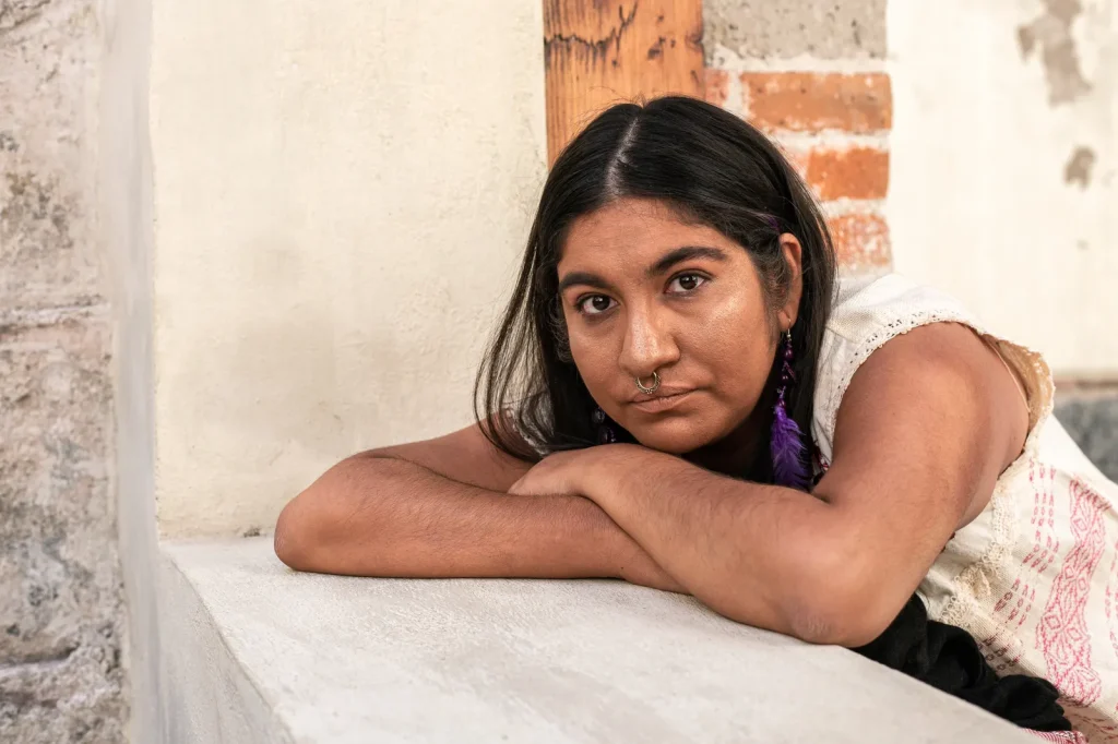 Mujer joven de cabello largo apoya los brazos en una mesa blanca y mira con calma