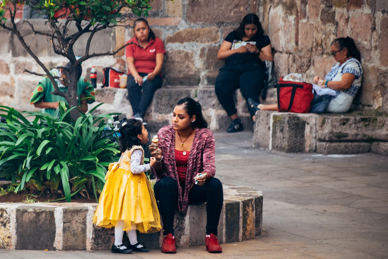 Mujer sentada en una plaza come un helado junto a una niña pequeña con vestido amarillo