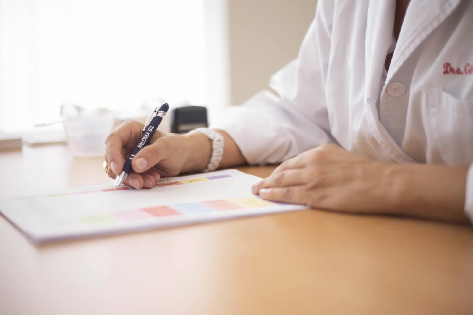 Manos de una médica con bata blanca escriben en una hoja sobre un escritorio
