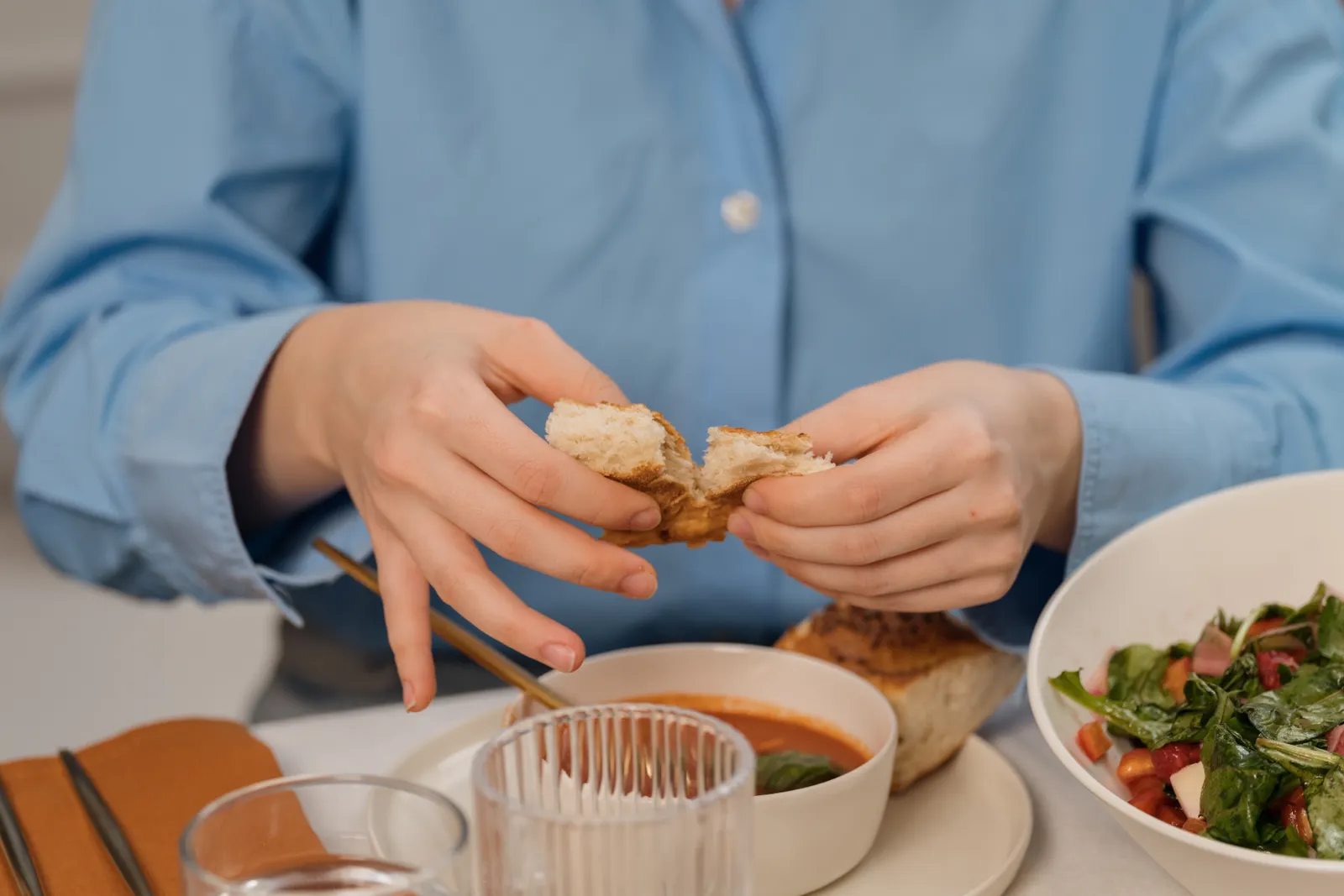 Manos de una mujer parten un pan sobre una mesa con sopa y ensalada