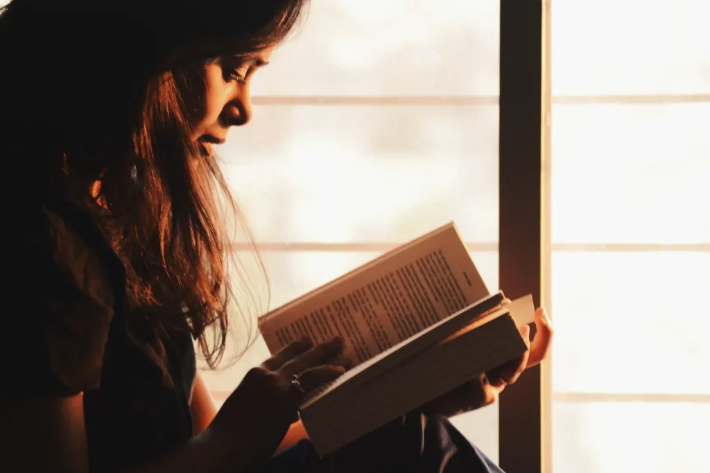 Mujer de cabello largo lee un libro junto a una ventana con luz cálida de la tarde