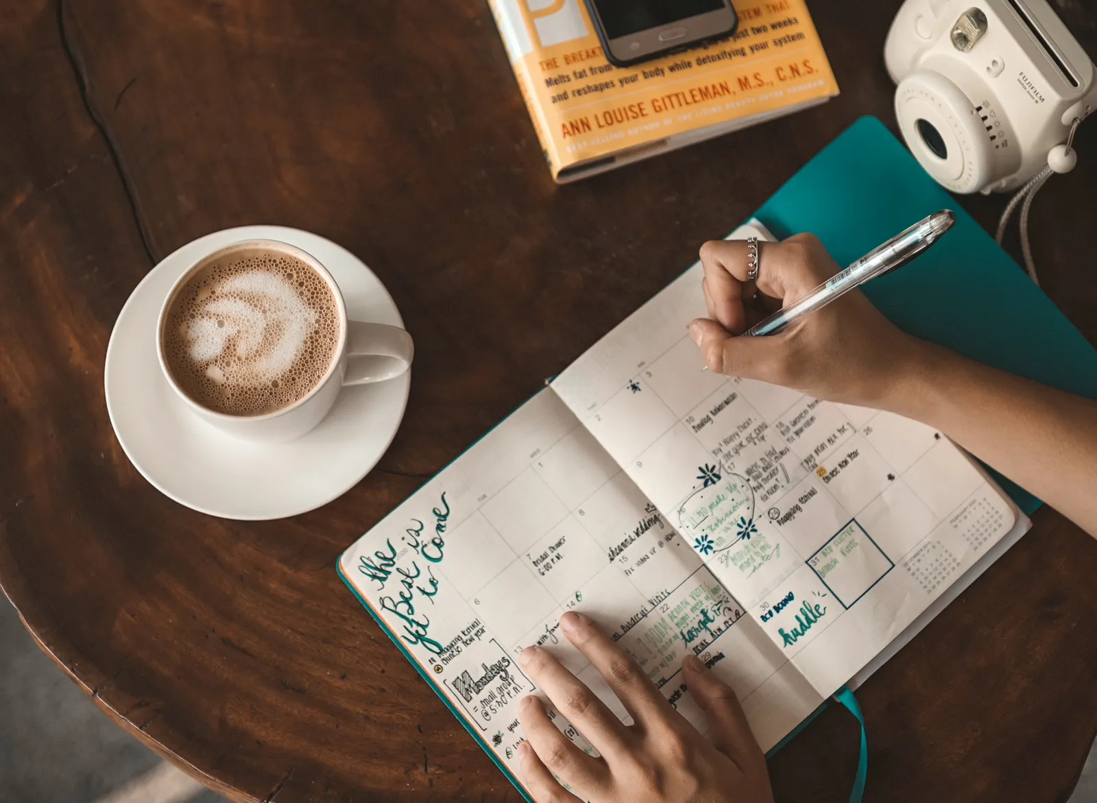 Manos de mujer escriben en una agenda abierta junto a un capuchino sobre una mesa de madera