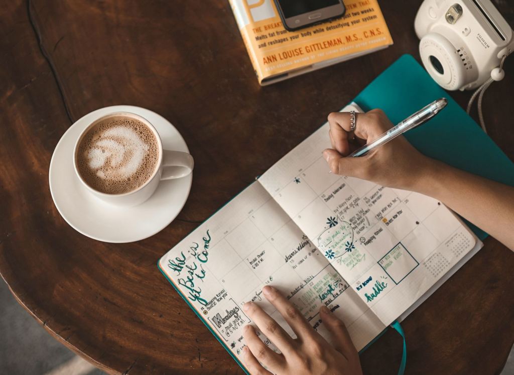 Mujer escribe en su agenda junto a un capuchino y un libro sobre una mesa de madera.