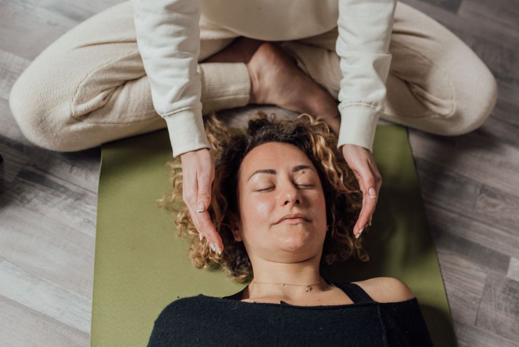 Mujer vestida y recostada sobre un tapete recibe una sesión de reiki, con las manos de la terapeuta cerca de su cabeza.
