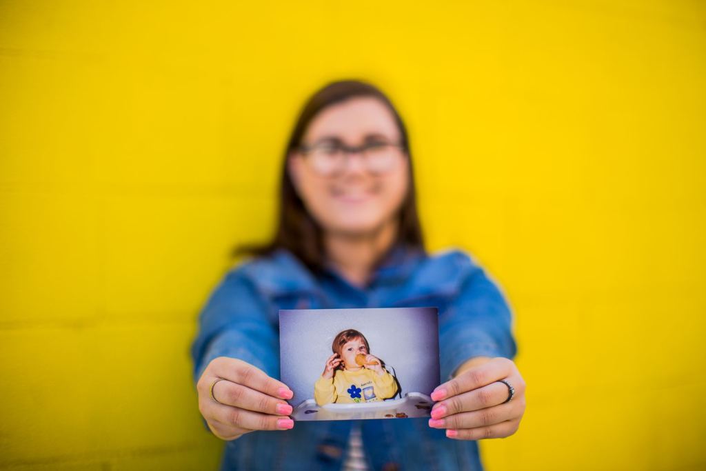 Mujer sonriente sostiene una foto de su infancia frente a un fondo amarillo.
