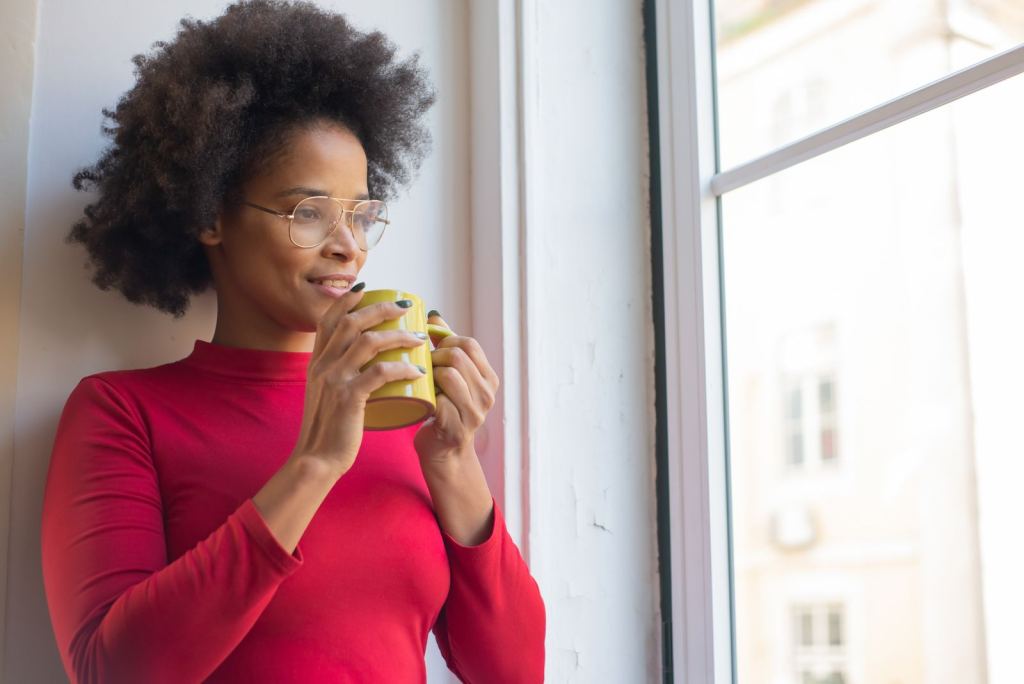Mujer con suéter rojo sostiene una taza y mira por la ventana con calma.