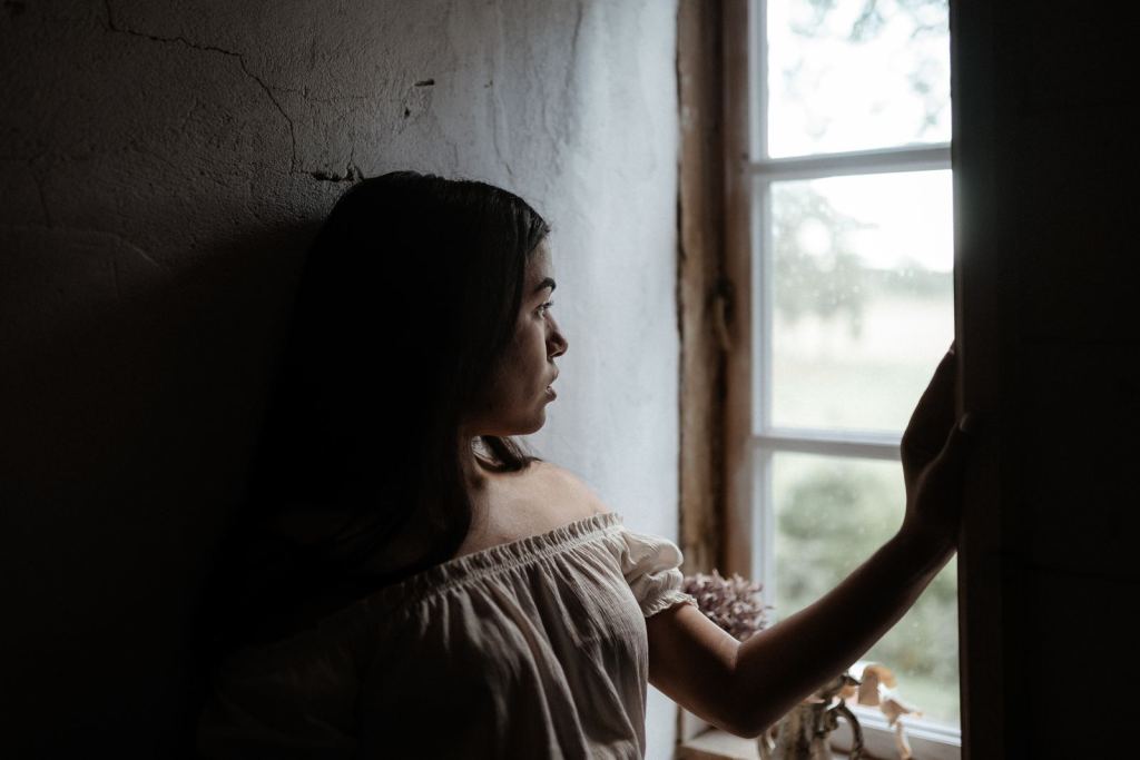 Mujer mira por la ventana en una habitación con luz y sombra.